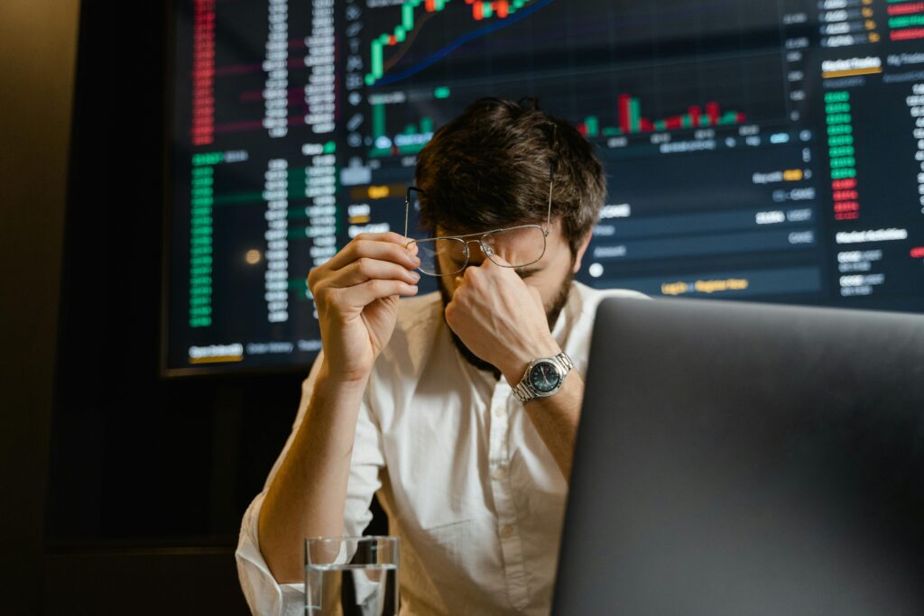 Frustrated man removing glasses in front of computer and data screens, overwhelmed by manual tasks and information overload symbolizing MSP burnout and the need for automation.