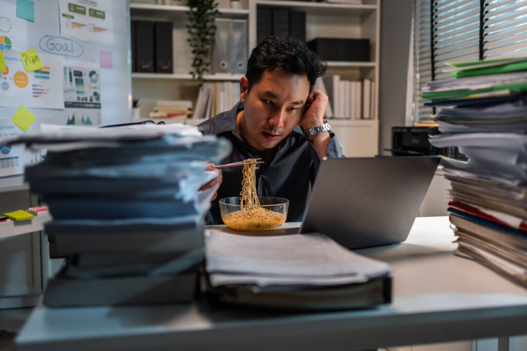 Asian young businessman eating noodles while working in office at night. Attractive professional male employee sitting on table, feeling hungry and starving then having dinner in corporate workplace.